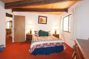 Un dormitorio con techo de madera, una cama con colcha estampada, dos lámparas de noche, una ventana con cortinas y una puerta que da a otra habitación.