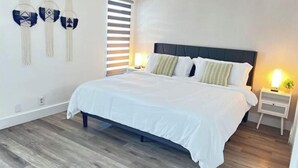 A modern bedroom with a large bed, two bedside tables with lamps, a wall-mounted light fixture, and a window with blinds. - Amazing bohemian house with a pool in a very nice neighborhood  (Sunrise)