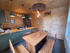 Interior de una cabaña de madera con cocina, mesa de comedor, bancos, lámparas colgantes y una pared de piedra.