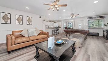 A spacious living room with a brown leather sofa, a dark wooden coffee table, and a pool table.