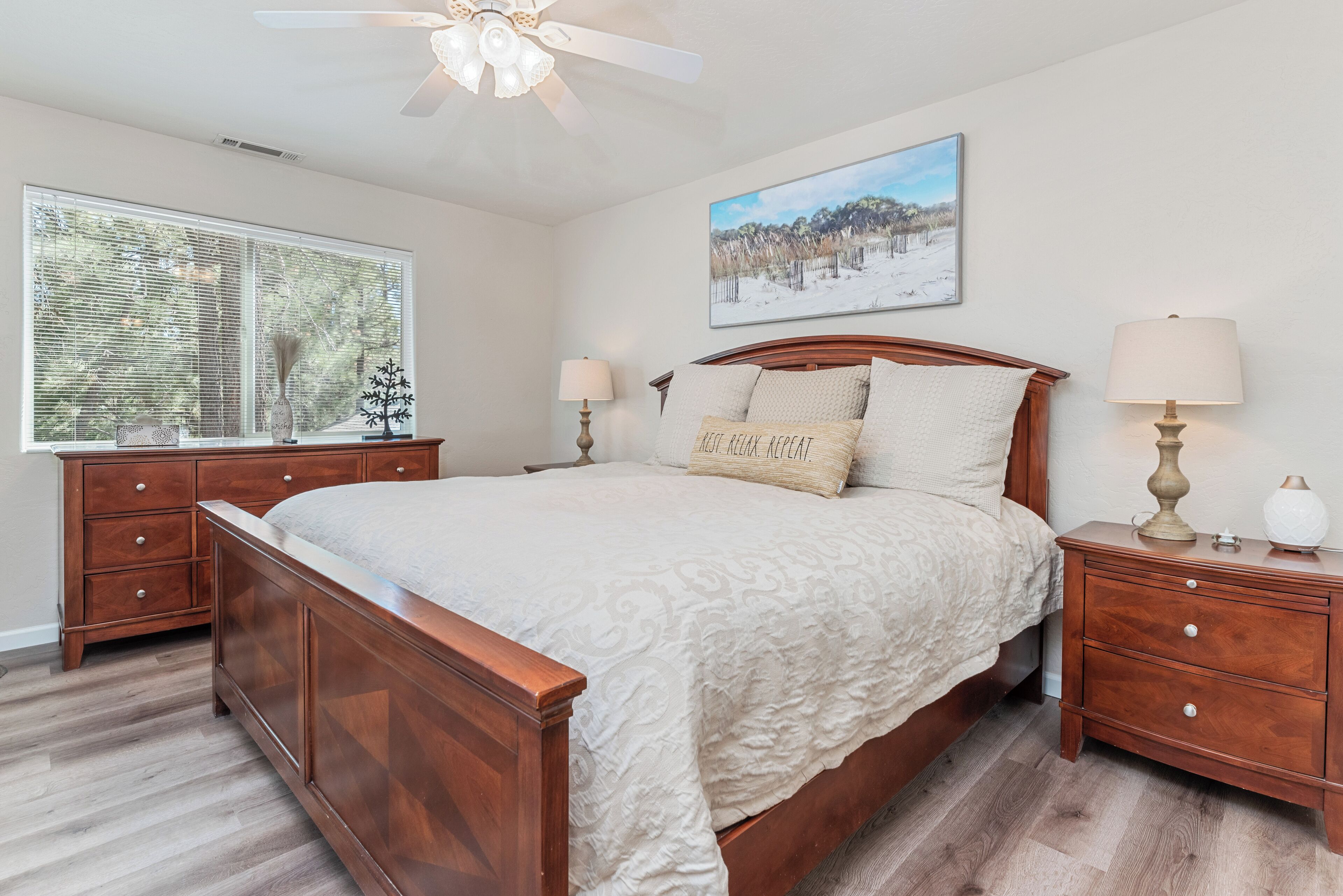 A bedroom with a wooden bed, two bedside tables, a nightstand, a ceiling fan, a window with blinds, and a framed picture on the wall.