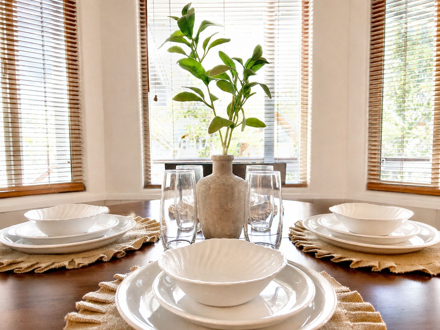 Spisebord dækket med hvide tallerkener, skåle og glas, med en vase og plante i midten, og udsigt til vinduer med persienner på hver side.
