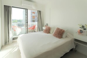 Habitación de hotel con una cama grande, un balcón con una silla roja y un escritorio blanco con una lámpara.
