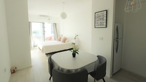 Ein modernes, helles Zimmer mit einem Bett, einem Esstisch mit Stühlen, einem Kühlschrank und einer Wandleuchte.