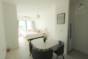 Et moderne, lyst værelse med seng, spisebord med stole, køleskab og vægmonteret lysarmatur.