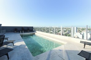 Piscina en la azotea con agua cristalina, rodeada de sillas de descanso y vistas a la ciudad.