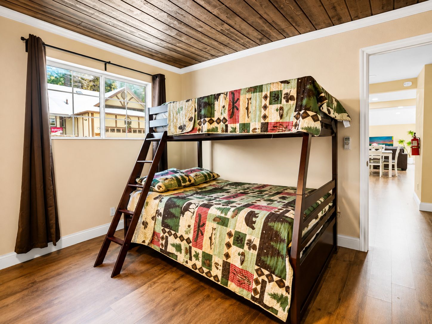 A bunk bed with a quilt, a wooden ladder, a window with curtains, and a view of a house outside.