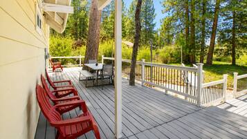 Una terraza de madera con mobiliario exterior, barandilla blanca y vista a un bosque.