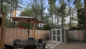 A wooden deck with a round table and chairs, a small shed, and a wooden fence. - Shady Pine Cottage (Big Bear City)