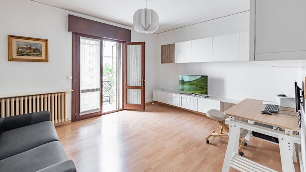 A modern living room with a sofa, a TV, a desk with a computer, and a sliding glass door leading to a balcony.