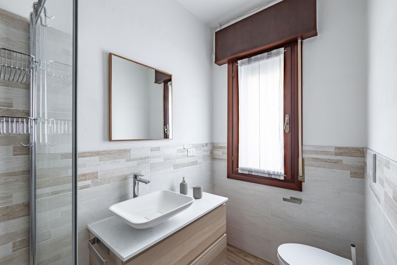 A modern bathroom with a glass shower enclosure, a white sink, and a window with a wooden frame.