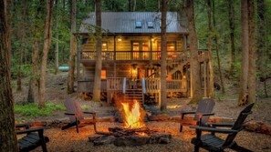 A cabin with a fire pit, two chairs, and a staircase leading to a deck. - Water's Edge - Pet Friendly | Creek Front | Hot Tub | Fire Pit (Cherry Log)