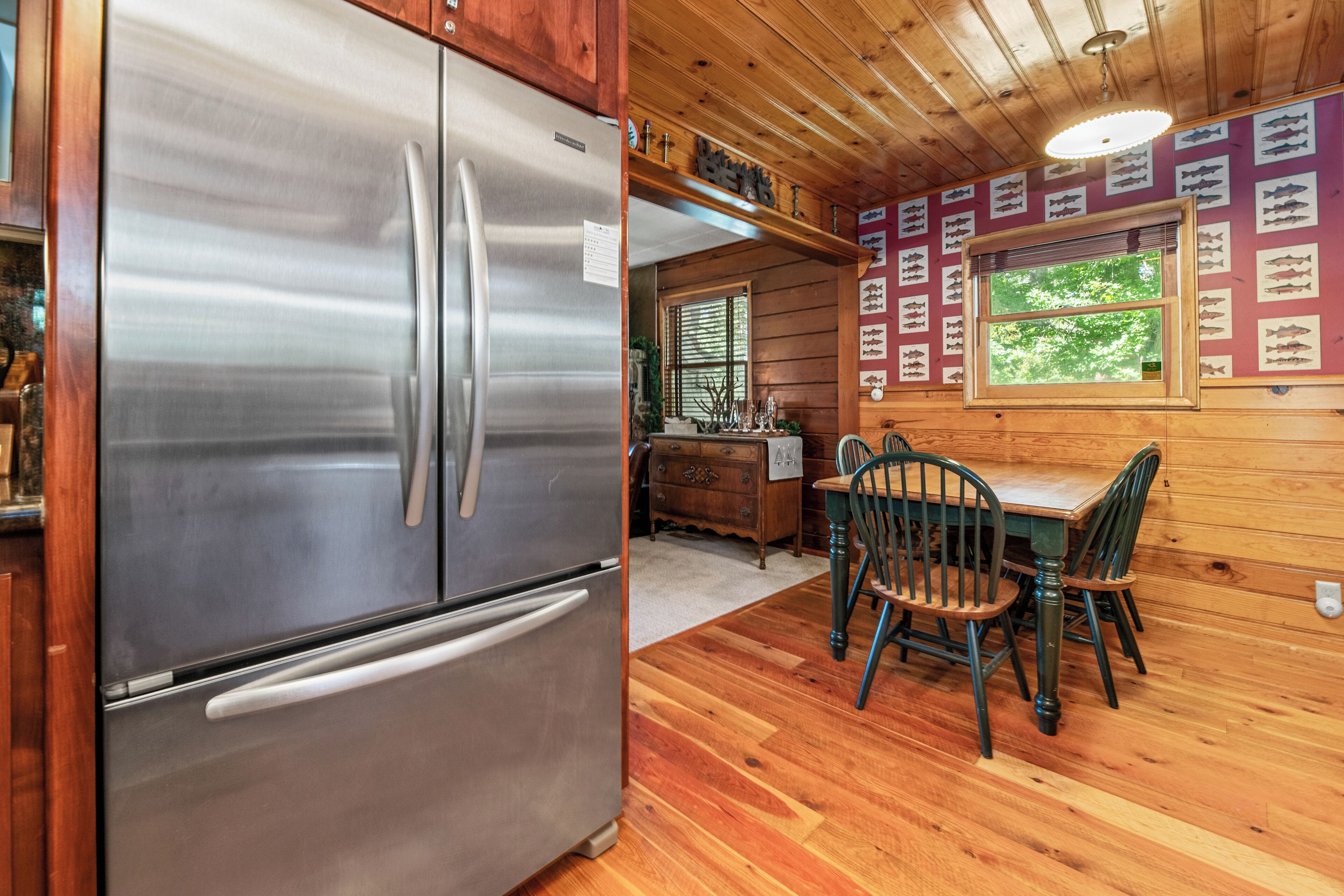A modern kitchen with a stainless steel refrigerator, wooden dining table with chairs, and a window with a view of greenery.