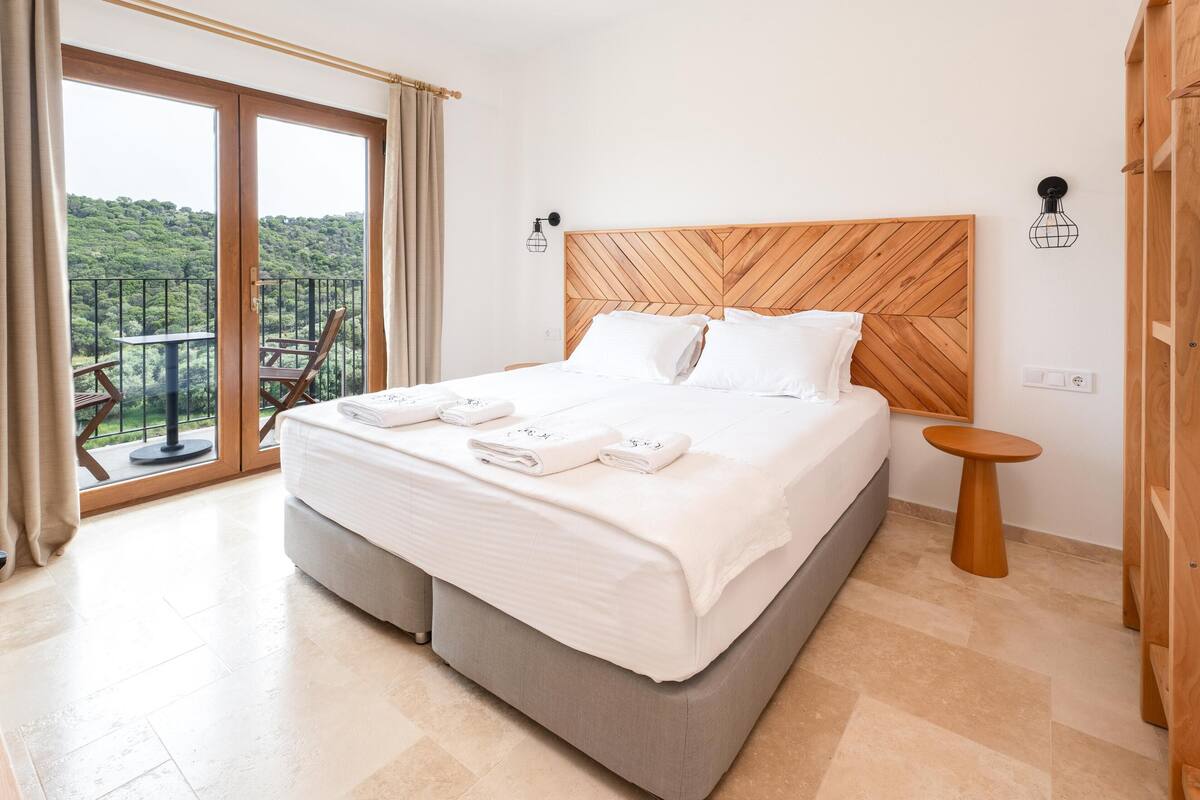A hotel room with a large bed, a wooden headboard, a balcony with a view of the forest, and a small round table.