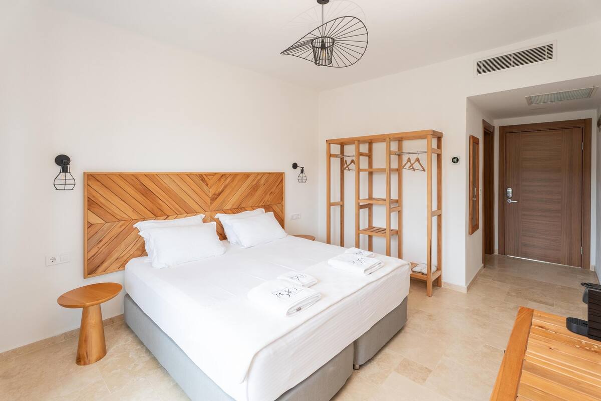 A modern hotel room with a wooden headboard, a bed with white linens, a small wooden side table, and a wooden wardrobe.