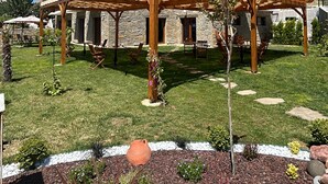 Una casa de dos pisos con fachada de piedra, una pérgola de madera y un jardín cuidado con diversas plantas y un pequeño árbol.