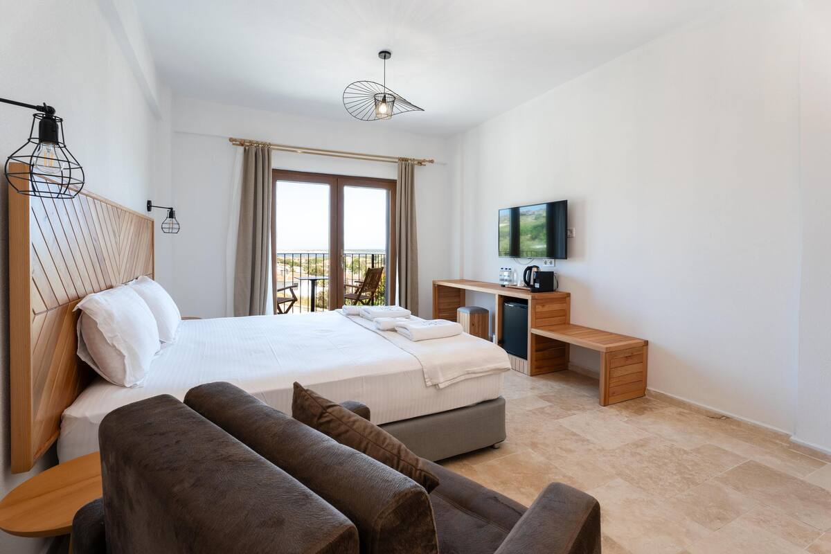 A modern hotel room with a large bed, a desk, a television, and a balcony with a view.