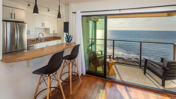 Cozinha moderna com bancada de madeira, eletrodomésticos de aço inoxidável e uma grande porta de vidro deslizante que dá para uma varanda com vista para o mar.