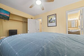 A bedroom with a bed, a ceiling fan, a door, a framed picture, and a bathroom visible through an open door. - Irie's Retreat (Sugarloaf)