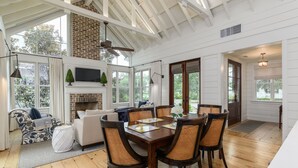 Una habitación amplia con una mesa de comedor rodeada de sillas de mimbre, una chimenea y ventanas grandes que ofrecen vistas del exterior.