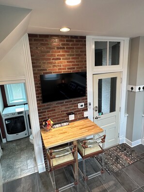 Compact kitchen with brick accent wall, flat-screen TV, oven, and two cushioned chairs.