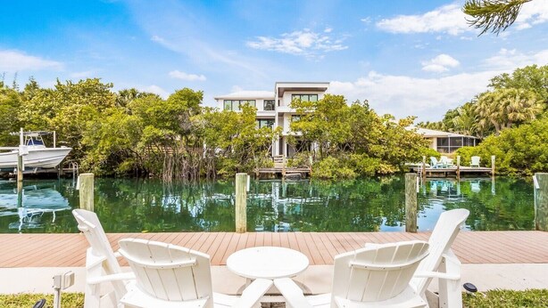 A waterfront view with a modern building, a dock with a boat, and a deck with chairs and a table. - Canal Place: Boat Dock, Pool & Spa, On Canal by Anna Maria Life Vacation Rentals (Anna Maria)