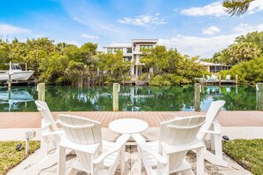 A waterfront view with a modern building, a dock with a boat, and a deck with chairs and a table. - Canal Place: Boat Dock, Pool & Spa, On Canal by Anna Maria Life Vacation Rentals (Anna Maria)
