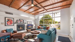 A spacious living room with a fireplace, a large window with a view of greenery, and a ceiling fan. - Canal Place: Boat Dock, Pool & Spa, On Canal by Anna Maria Life Vacation Rentals (Anna Maria)