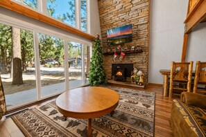 A cozy living room with a stone fireplace, a television mounted on the wall, and a large window overlooking a wooded area. - Sherwood Pines (Big Bear City)
