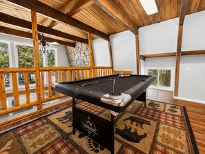 A pool table in a room with wooden beams, a stone wall, and a view through large windows. - Sherwood Pines (Big Bear City)