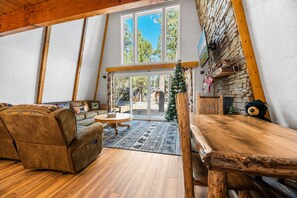 A cozy loft-style living space with a wooden dining table, a sofa, and a large window overlooking a natural landscape. - Sherwood Pines (Big Bear City)