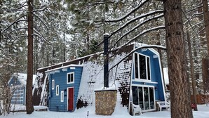 En snötäckt stuga med en skorsten av sten, en röd dörr och ett stort fönster.