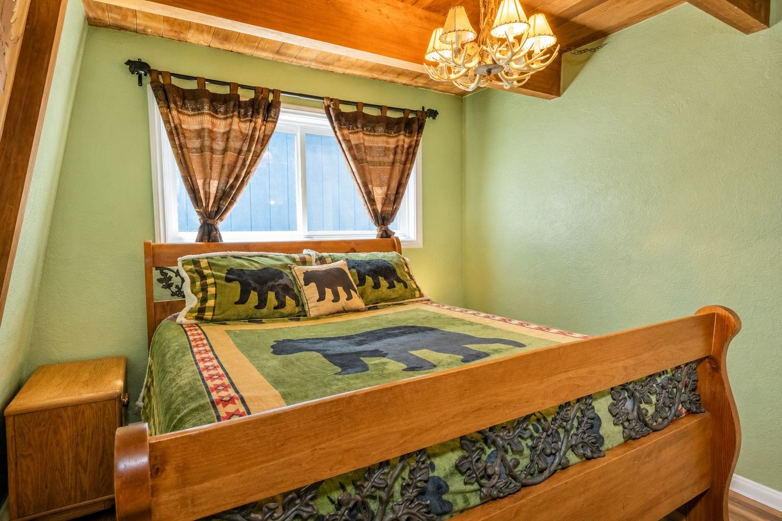 A wooden bed with a bear-themed bedspread, a nightstand, a window with curtains, and a chandelier.