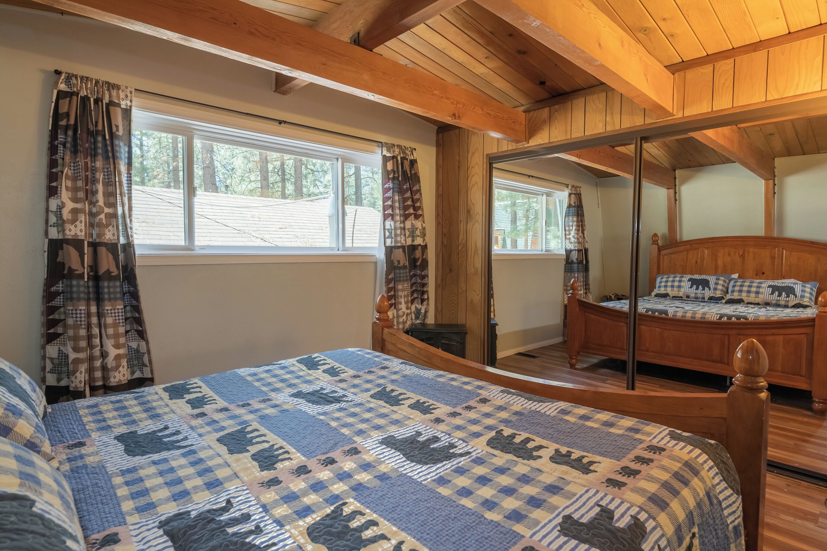 Un dormitorio con techo de madera, una cama grande con colcha a cuadros, dos ventanas con cortinas y un espejo en la pared.