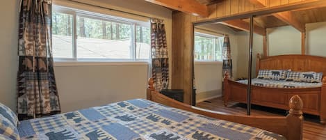 Un dormitorio con techo de madera, una cama grande con colcha a cuadros, dos ventanas con cortinas y un espejo en la pared.
