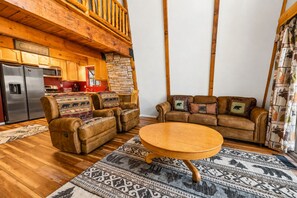 L’intérieur chaleureux d’un chalet avec une table à manger en bois, des canapés en cuir à motifs animaliers, une cheminée en pierre et une cuisine équipée d’un réfrigérateur.