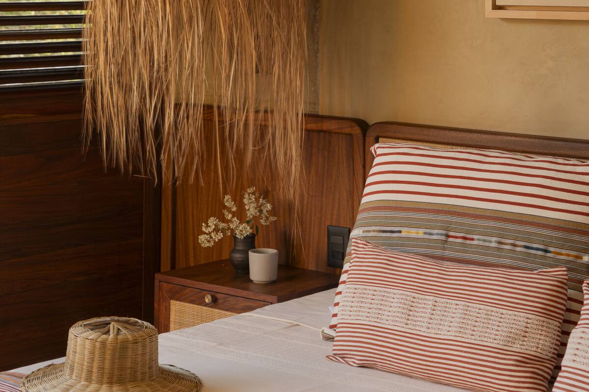 Una cama con ropa de cama a rayas, una lámpara de fibra y un pequeño florero con flores sobre una mesita de noche de madera.
