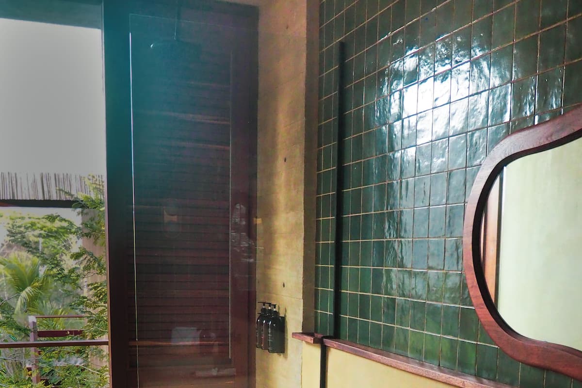 Un baño con un espejo grande, un lavabo color beige y un inodoro. Se ve vegetación a través de la ventana.