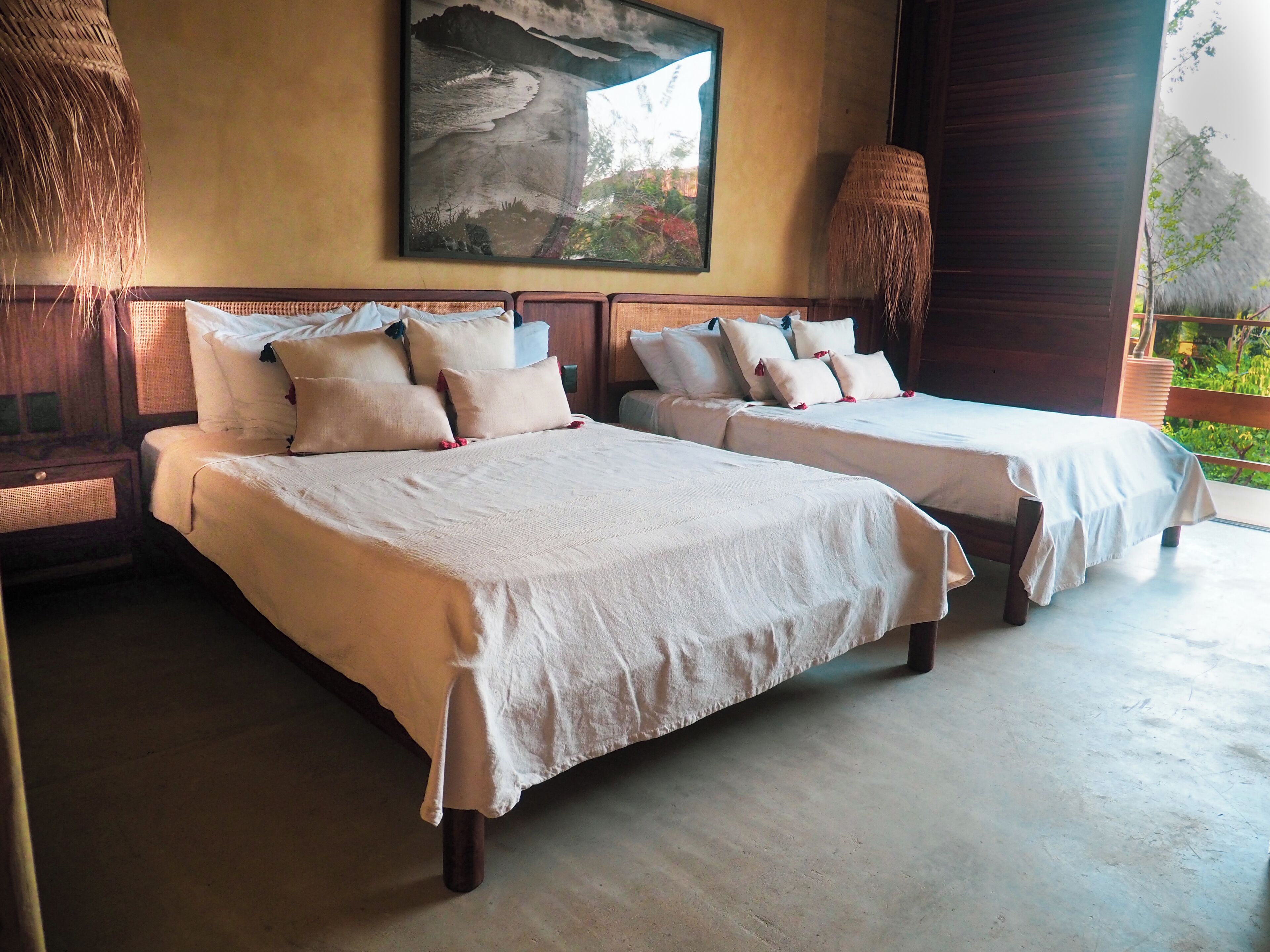 Dos camas bien hechas con sábanas blancas y varios almohadones, un cuadro grande en la pared y una cesta tejida al lado.