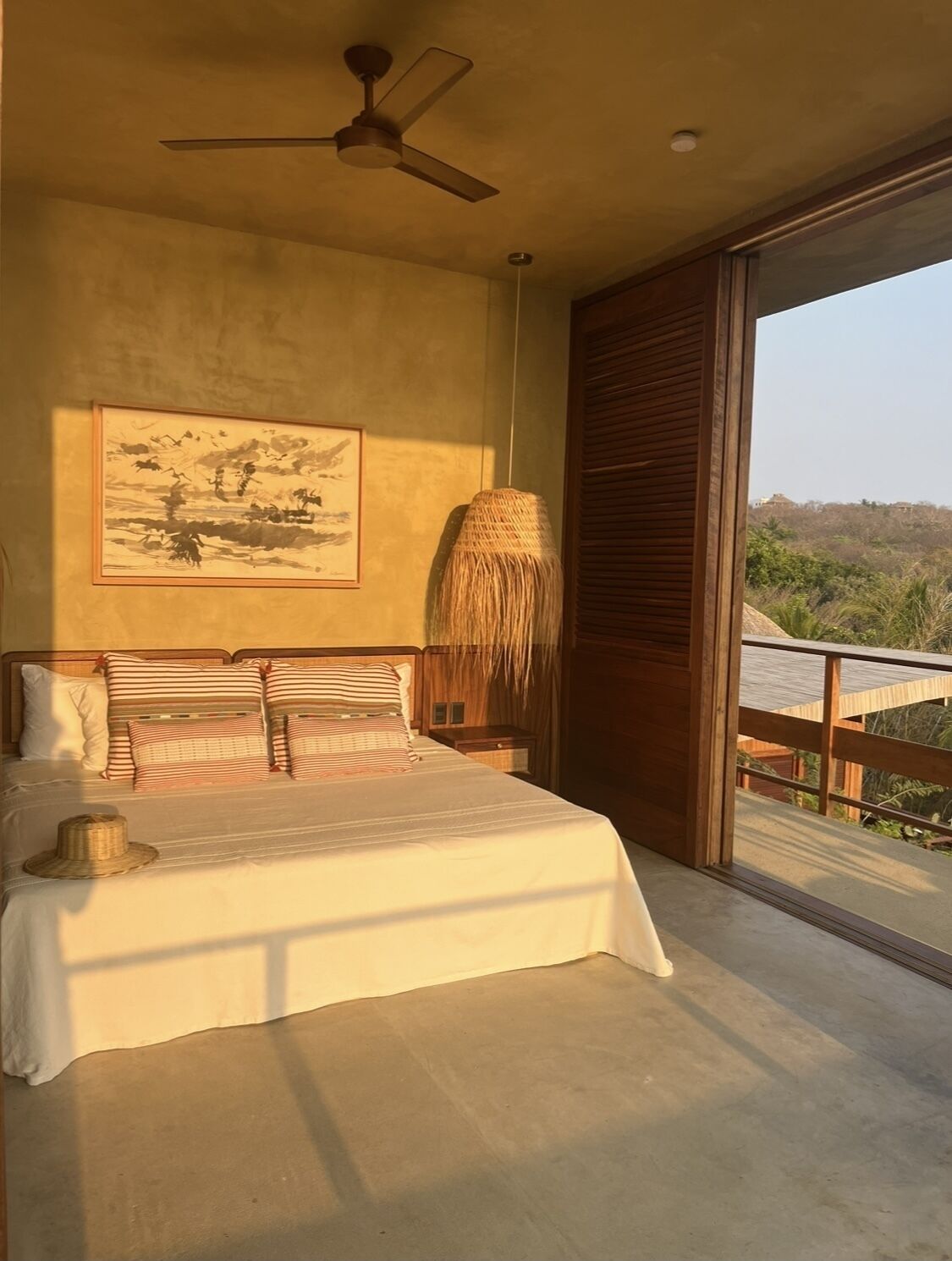 A bedroom with a bed, a ceiling fan, a large window, and a view of the outdoors.