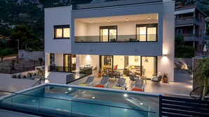 A modern two-story house with a swimming pool, outdoor seating, and a view of the mountains.