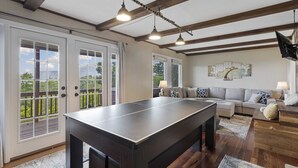 Un vaste espace de vie comprenant une table de ping-pong, un canapé et une table basse. De grandes fenêtres et un balcon avec vue sur la verdure complètent cet espace.