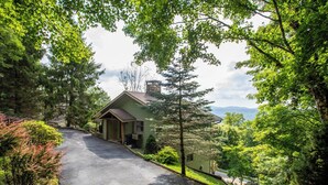 Une maison de plain-pied dotée d’un porche à l’avant, entourée d’arbres denses et offrant une vue dégagée sur une chaîne de montagnes en arrière-plan.