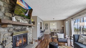 Un salon chaleureux avec une cheminée en pierre, un coin repas et une grande fenêtre donnant sur un paysage agréable.