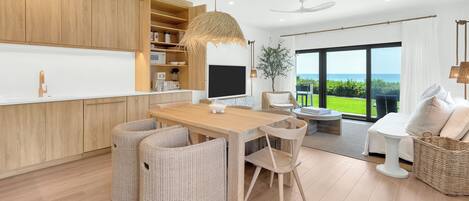 A modern kitchen with wooden cabinets, a dining area with wicker chairs, and a large window offering a view of the sea.