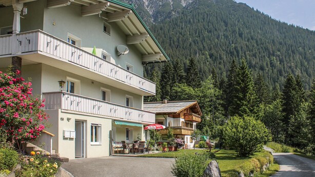 A two-story building with balconies, a restaurant area with outdoor seating, and a mountain backdrop. - Ferienwohnung - Wald am Arlb. (Wald am Arlberg)