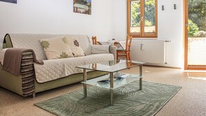 A living room with a sofa, a glass coffee table, a green rug, a dining table with chairs, and a window with blinds. - Ferienwohnung - Wald am Arlb. (Wald am Arlberg)
