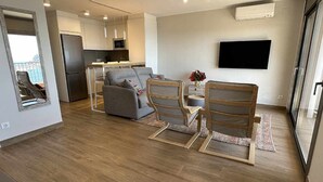 A modern living room with a grey sofa, wooden chairs, a dining table, and a kitchen area with a fridge and microwave.