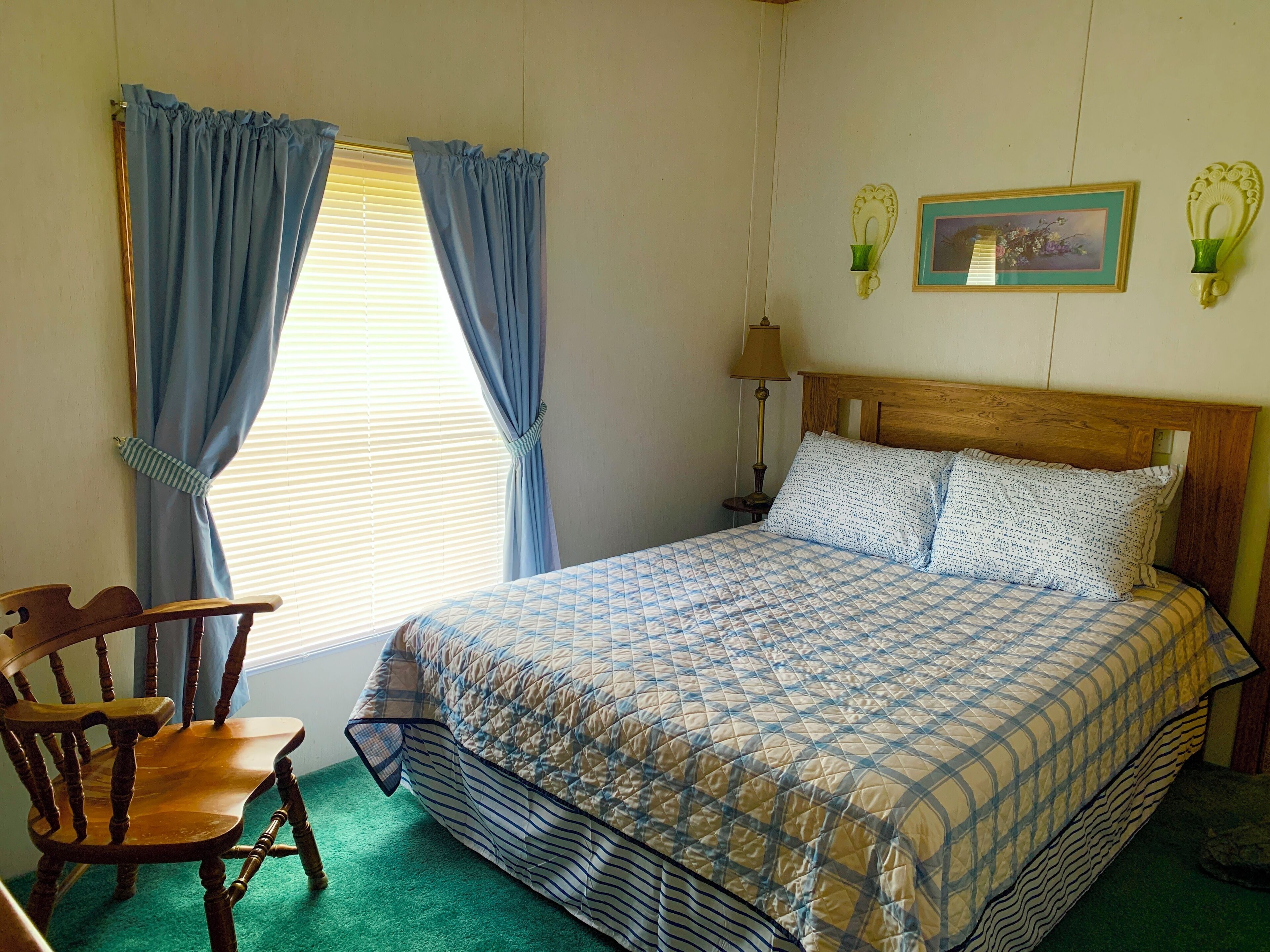 Un dormitorio con una cama de madera, una silla de madera, una alfombra verde y una ventana con cortinas.
