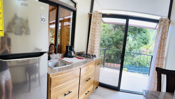 A compact kitchen with a refrigerator, sink, and wooden cabinets. A coffee maker and gas stove are on the countertop. The room has a sliding glass door leading to a balcony with a view of greenery. - Enchanted Monkey Apartment  (Quepos)